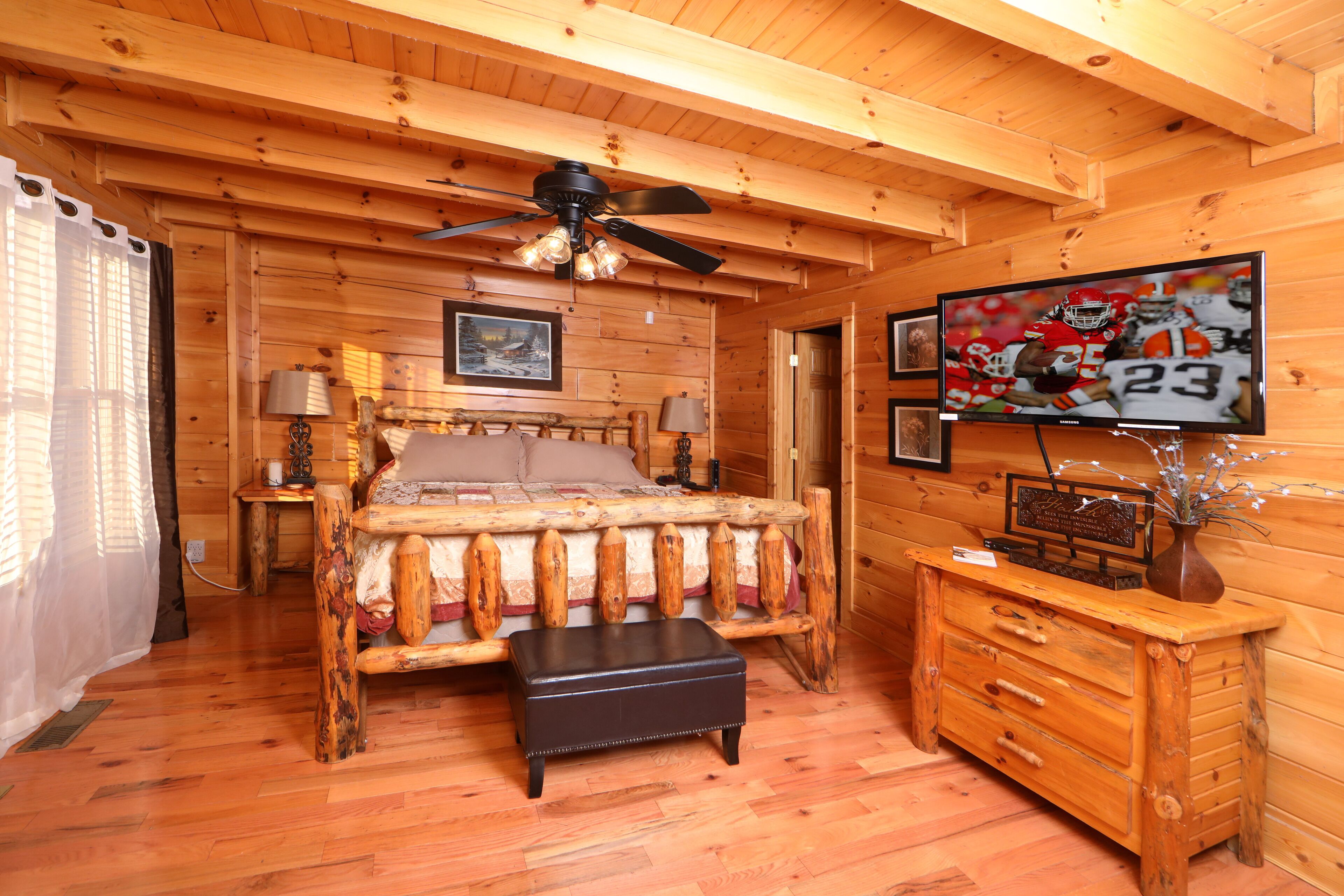 A wooden cabin bedroom with a bed, a dresser, a television, and a ceiling fan.