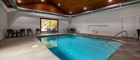 An indoor swimming pool with a wooden ceiling, stone flooring, and black chairs around the pool.