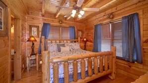 Ein Schlafzimmer in einer Holzhütte mit einem Bett, zwei Lampen, einem Deckenventilator und großen Fenstern mit Vorhängen.