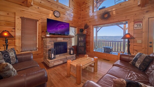 Ein gemütliches Blockhauss-Interieur mit Kamin aus Stein, Ledersofas, einem Flachbildfernseher und einem Balkon mit Aussicht.
