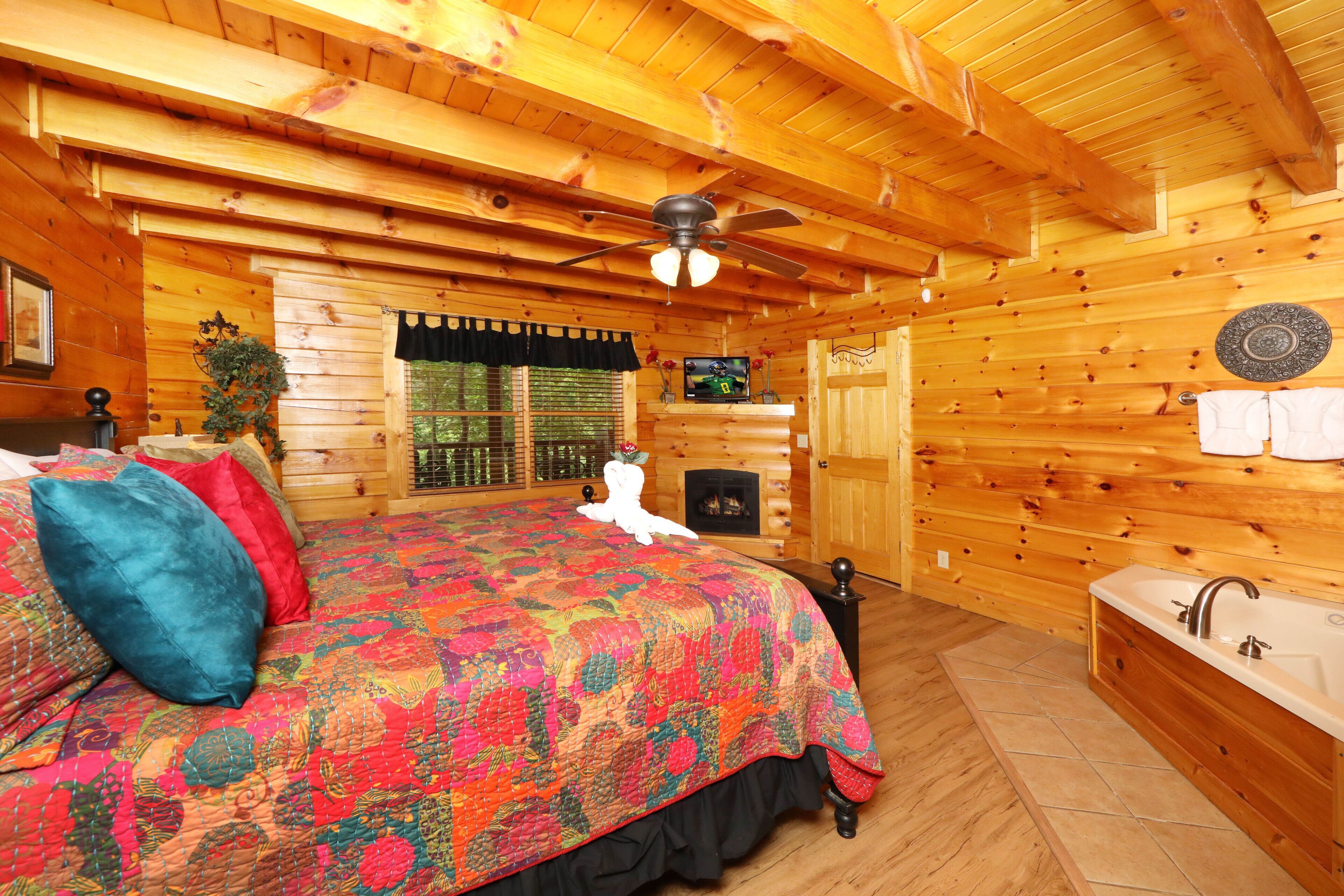 A log cabin bedroom with a large bed, a ceiling fan, a fireplace, and a wooden bathtub.