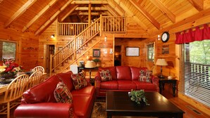 L’intérieur d’une cabane en rondins, avec un canapé en cuir rouge, une table à manger en bois et un escalier menant à un étage supérieur.