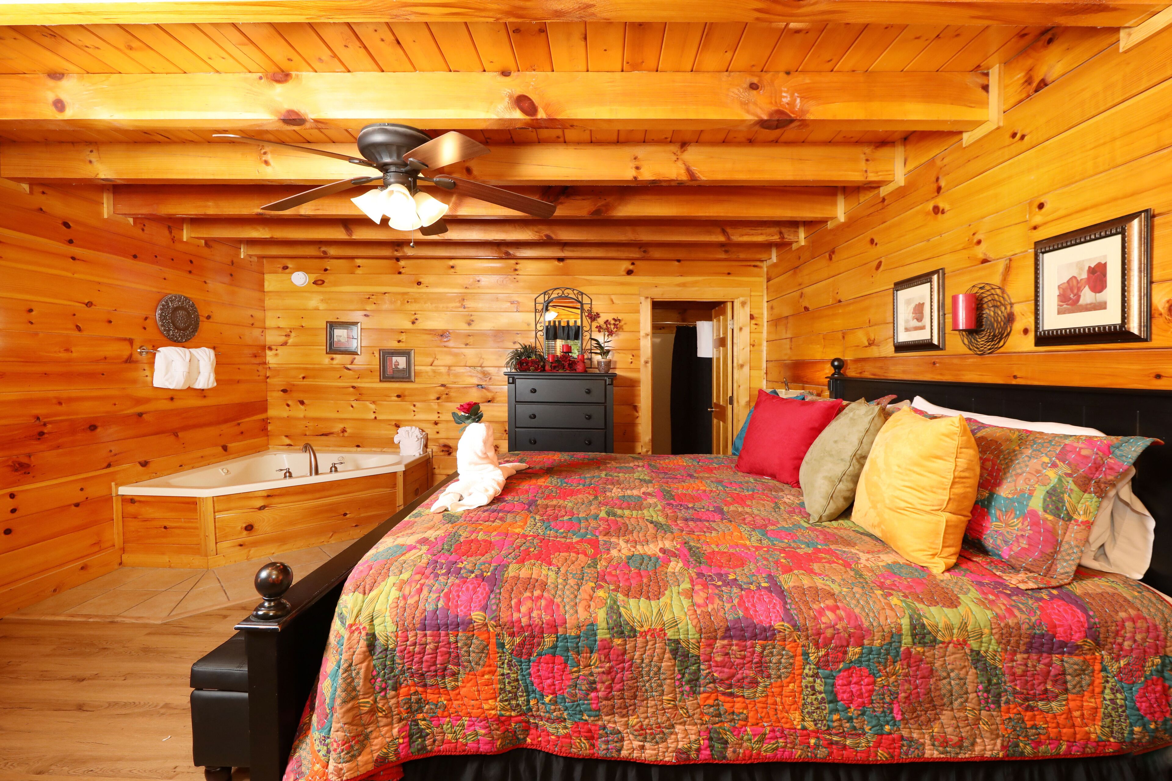A log cabin bedroom with a wooden ceiling, a bed with a colorful quilt, a dresser, and a vanity with a mirror.