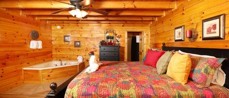A log cabin bedroom with a wooden ceiling, a bed with a colorful quilt, a dresser, and a vanity with a mirror.