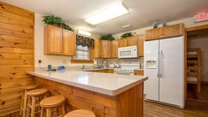 A kitchen with wooden cabinets, a white refrigerator, a microwave, and a bar area with stools. - Briarstone Lodge Condo 13F - Three Bedroom Condo (Pigeon Forge)