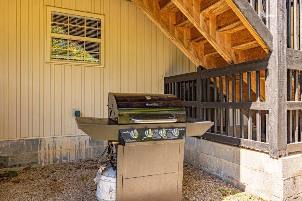 A charcoal grill with four burners, positioned outdoors next to a building with a wooden deck and a window with a screen.