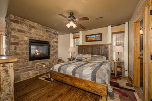 A bedroom with a stone fireplace, a bed with a quilt labeled "WILDWOOD," a ceiling fan, and a painting on the wall. - Hilltop Haven - Four Bedroom Chalet (Gatlinburg)
