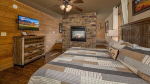 A bedroom with a stone fireplace, wooden walls, a bed with patterned bedding, a wooden dresser, and a ceiling fan. - Hilltop Haven - Four Bedroom Chalet (Gatlinburg)