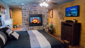 Ein Schlafzimmer mit Kamin aus Stein, einem Bett mit gestreifter Bettwäsche, einem Deckenventilator und einem an der Wand befestigten Fernseher.