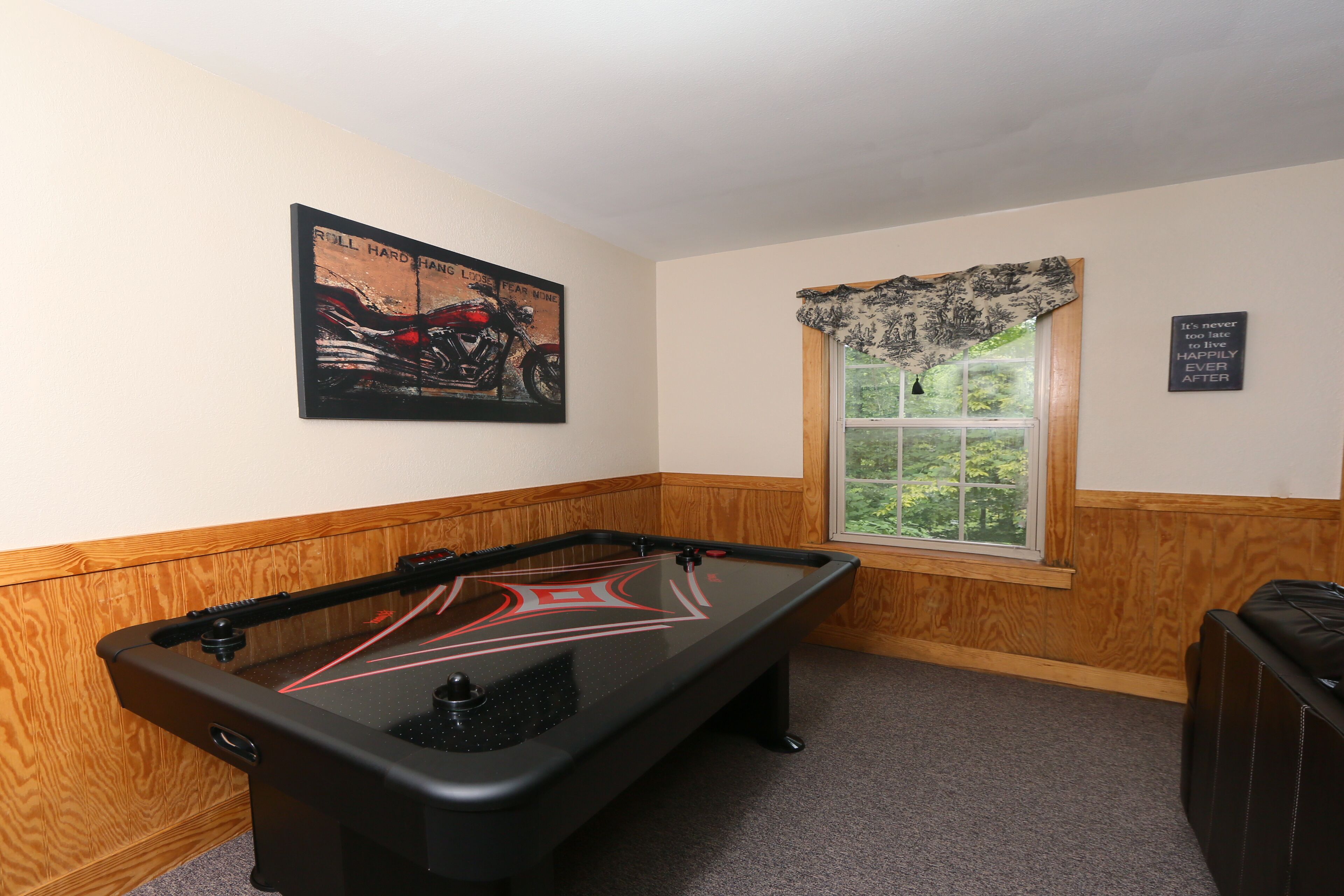 A pool table in a room with wooden paneling, a window with a view of greenery, and a poster on the wall.