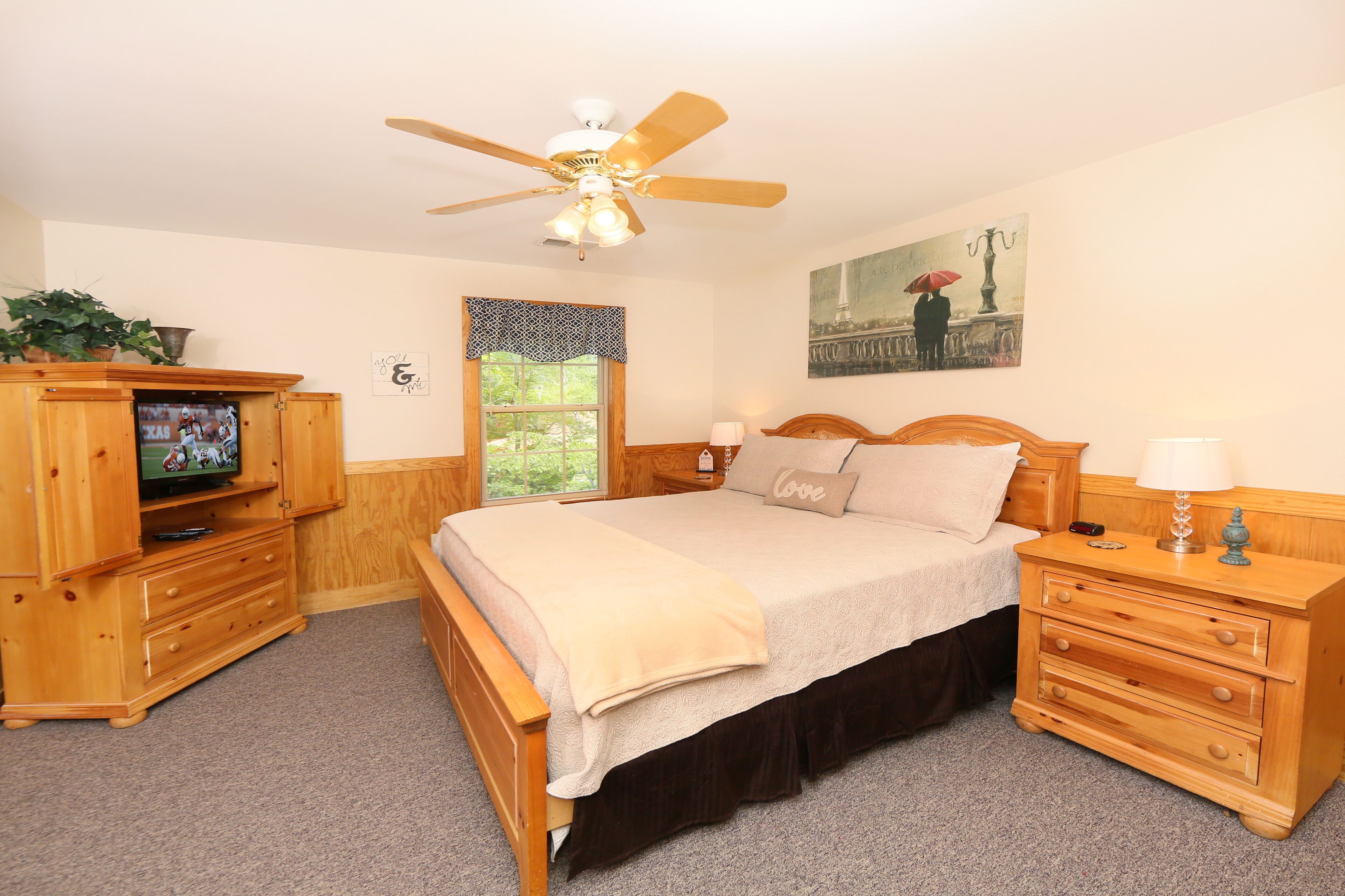 A bedroom with a bed, wooden nightstands, a TV, a ceiling fan, a window, and a painting.