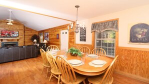A dining area with a wooden table set for six, surrounded by wooden chairs. A chandelier hangs from the ceiling, and a fireplace is visible in the background. - Holly Grove 1 - Three Bedroom Chalet (Pigeon Forge)