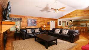 A cozy living room with a fireplace, a ceiling fan, and a kitchen in the background. - Holly Grove 1 - Three Bedroom Chalet (Pigeon Forge)