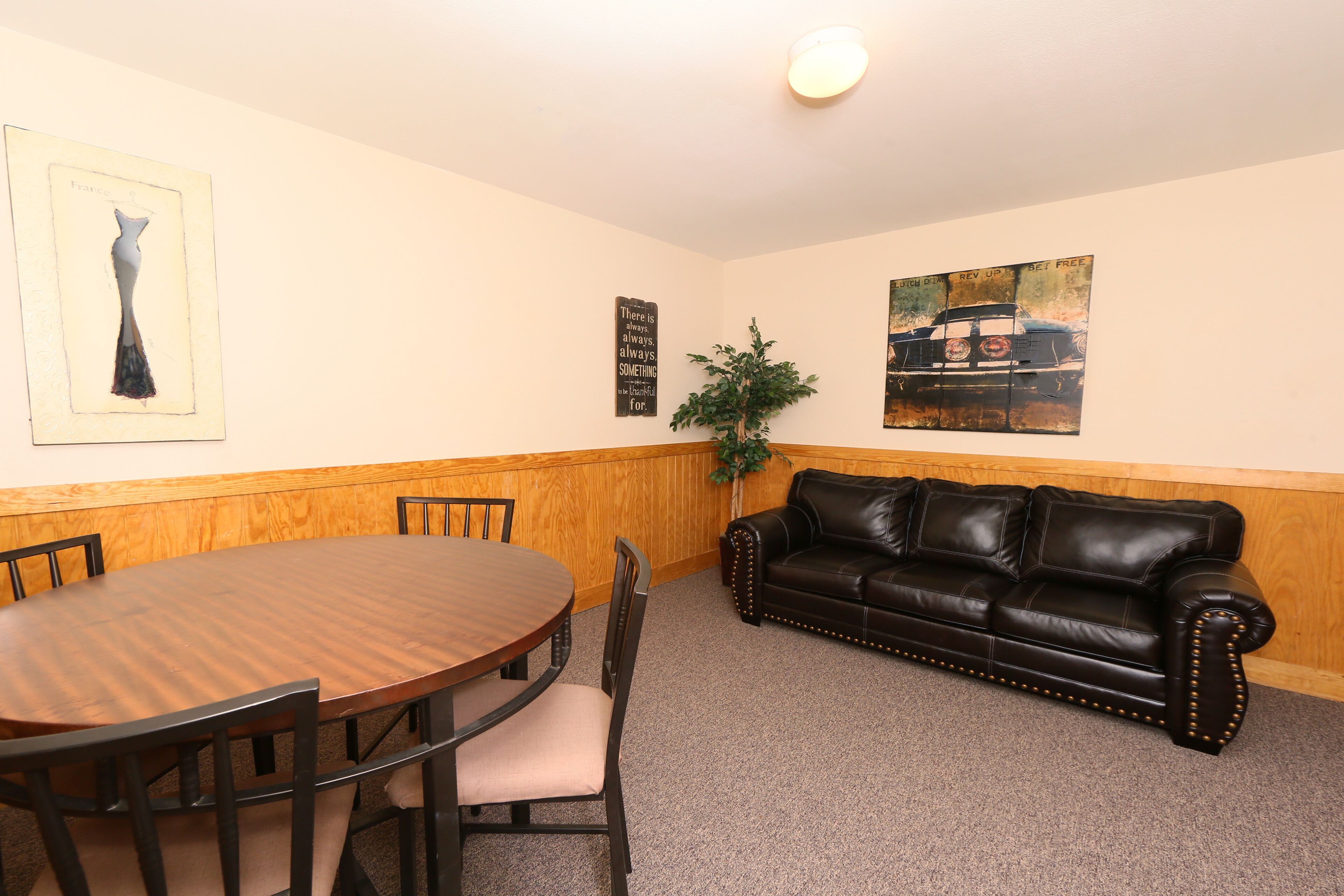A room with a wooden dining table and chairs, a black leather sofa, a potted plant, and artwork on the walls.