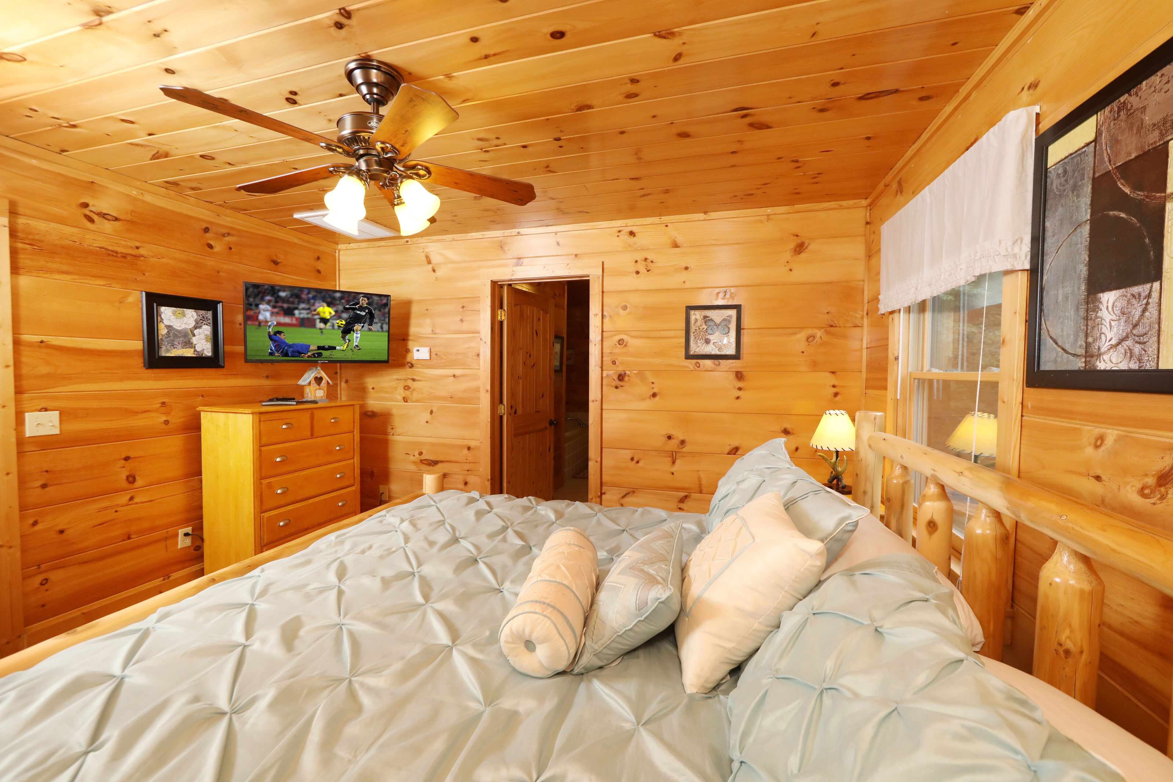 A wooden cabin bedroom with a bed, ceiling fan, dresser, and framed pictures on the wall.