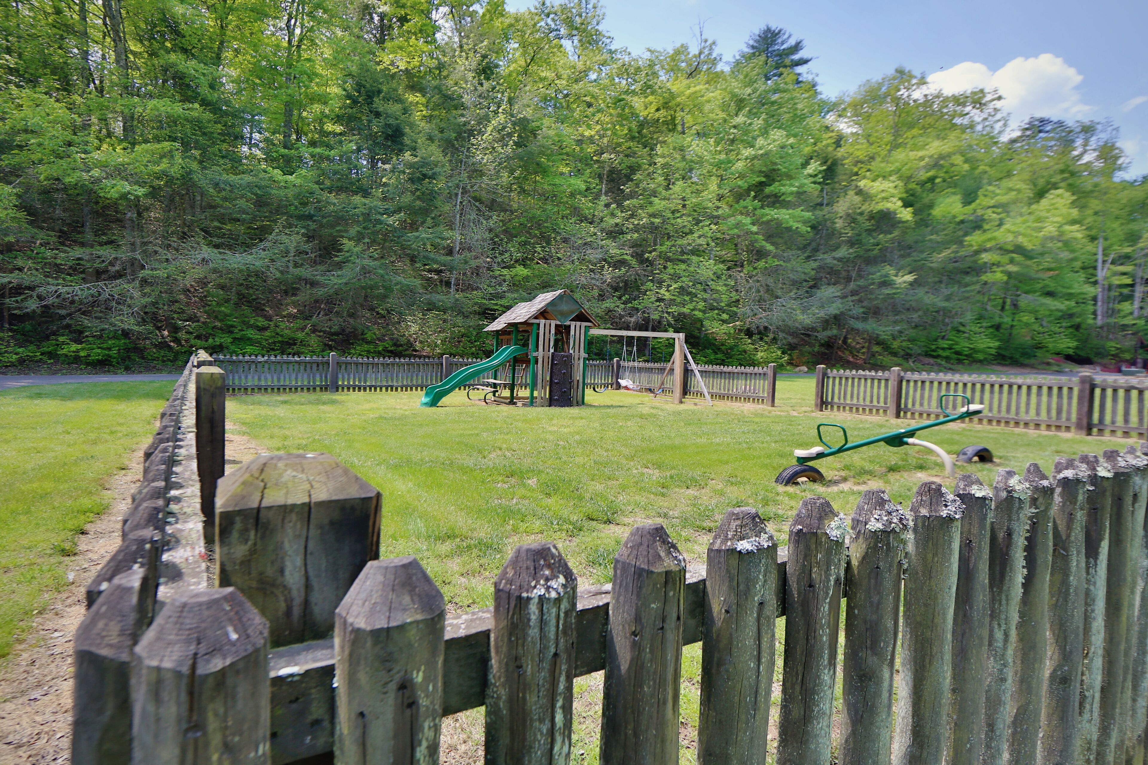 A playground with a slide and swings, surrounded by a wooden fence and trees.