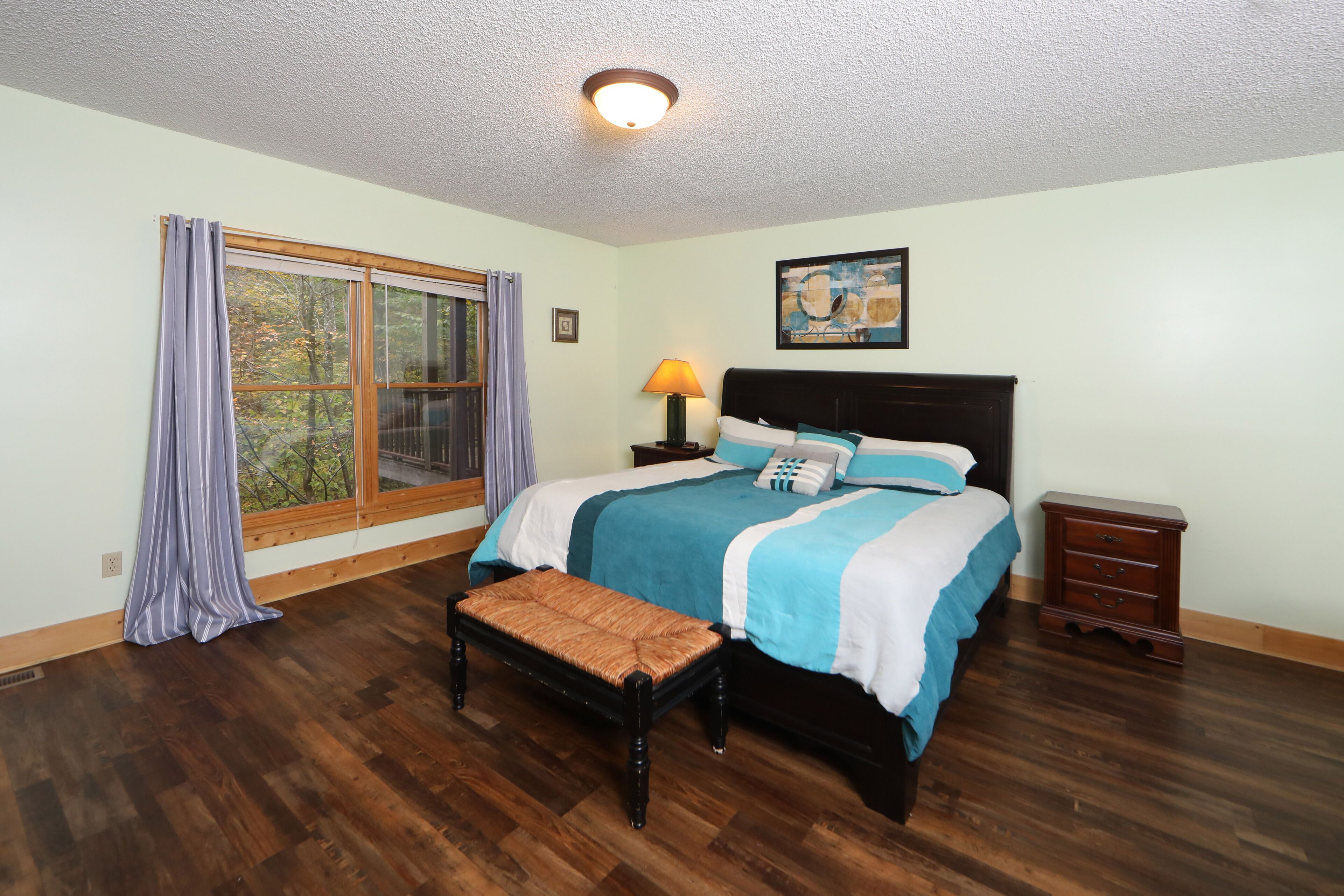 A bedroom with a large window, a bed with a headboard, a bedside table with a lamp, and a wooden bench.
