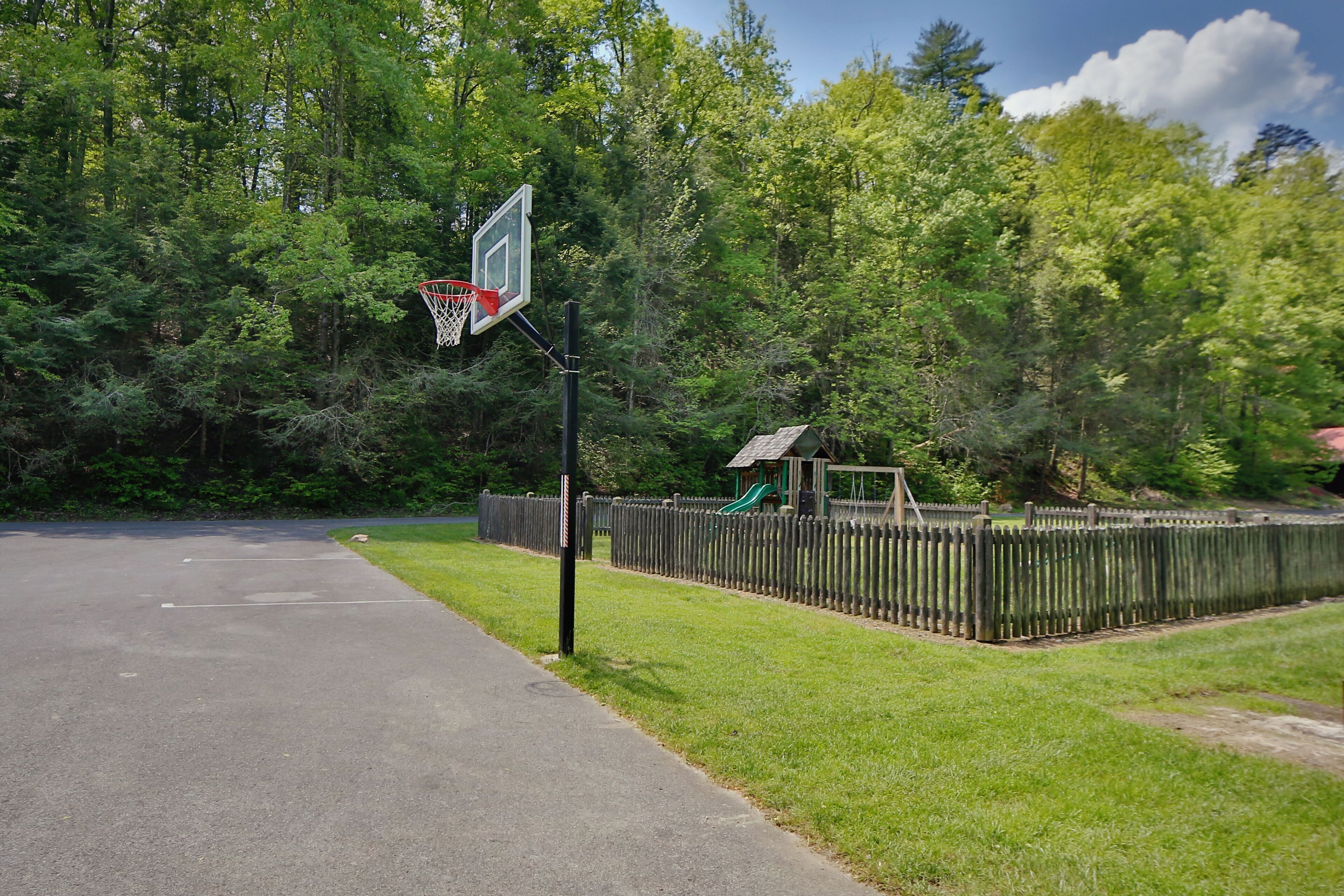 Un terrain de basket-ball entouré d’une clôture en bois, avec une aire de jeux et une zone boisée en arrière-plan.