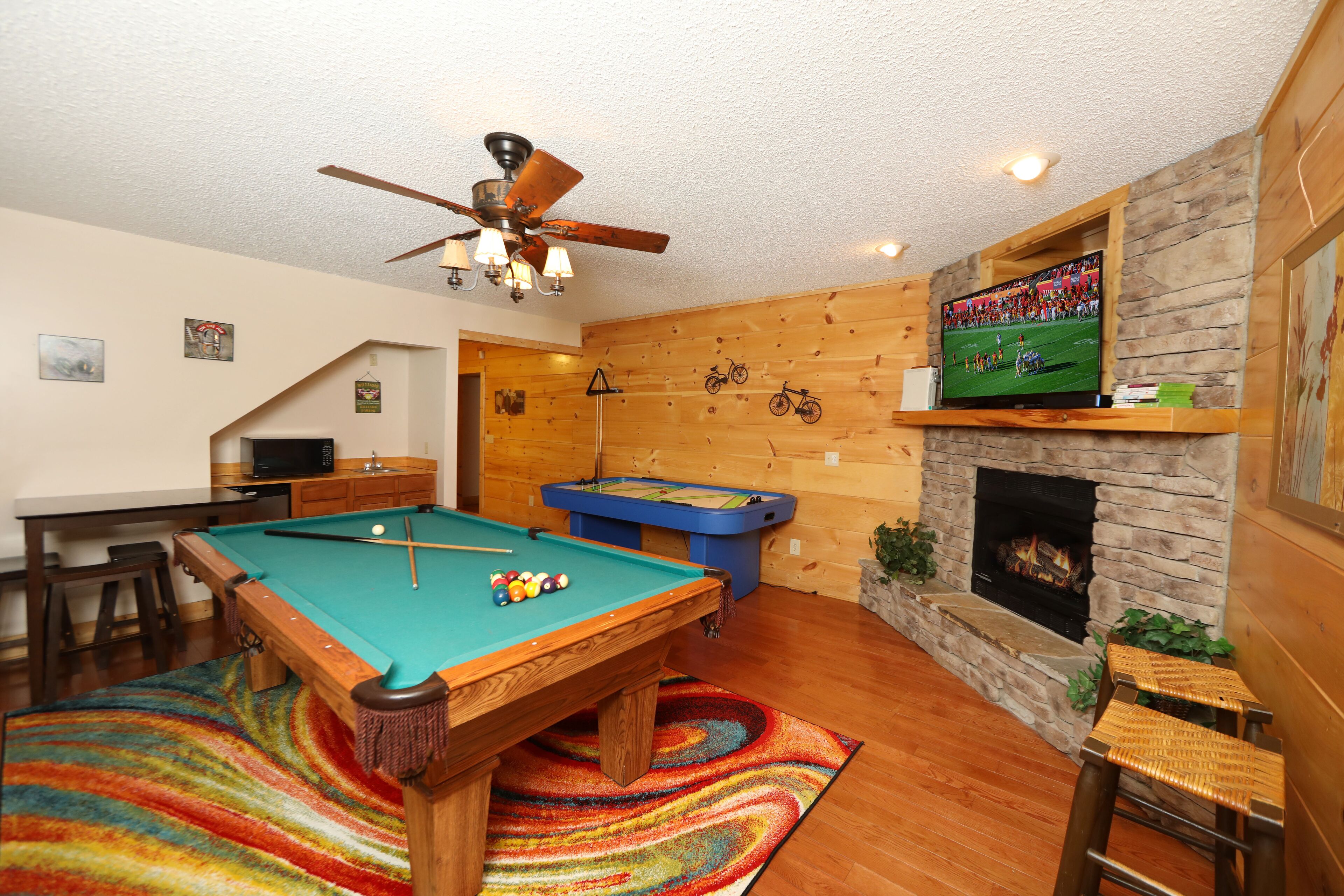 A pool table, a foosball table, a television, and a fireplace in a room with wooden walls and a ceiling fan.