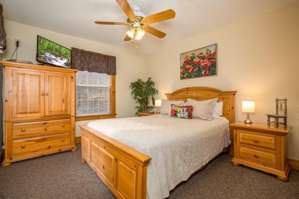 Une chambre à coucher avec un lit en bois, un ventilateur de plafond, un téléviseur posé sur un meuble et une fenêtre avec des stores.