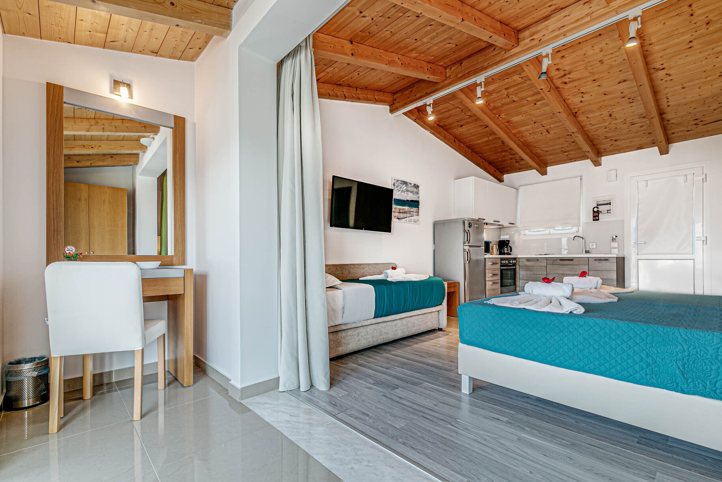 A modern bedroom with a wooden ceiling, a bed with teal bedding, a desk with a chair, and a television on the wall.