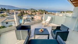 A balcony with wicker furniture, a small table with fruit, and a view of a coastal town and mountains.