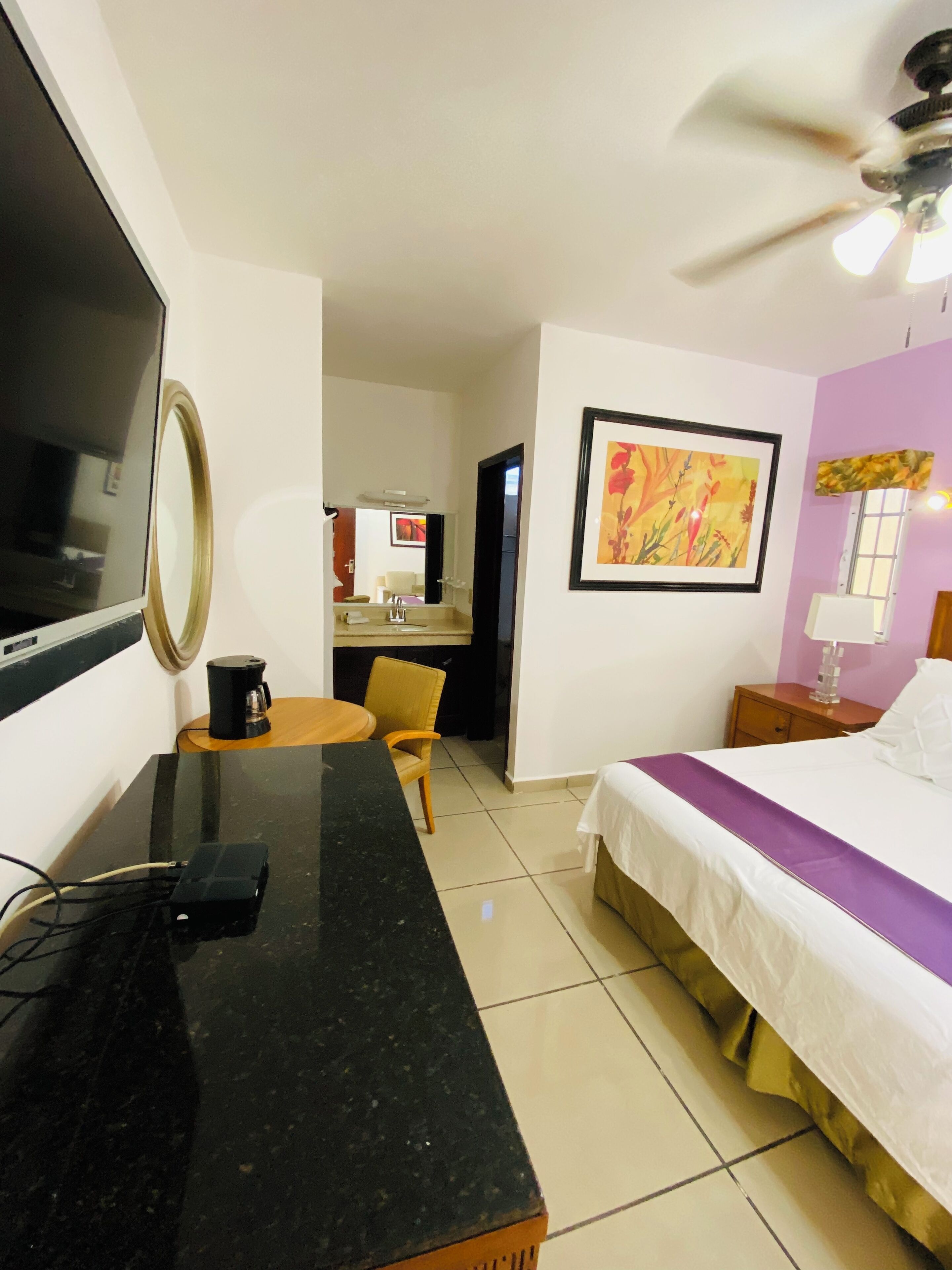 A hotel room with a bed, a desk with a coffee maker, a chair, a TV, a painting, and a ceiling fan.