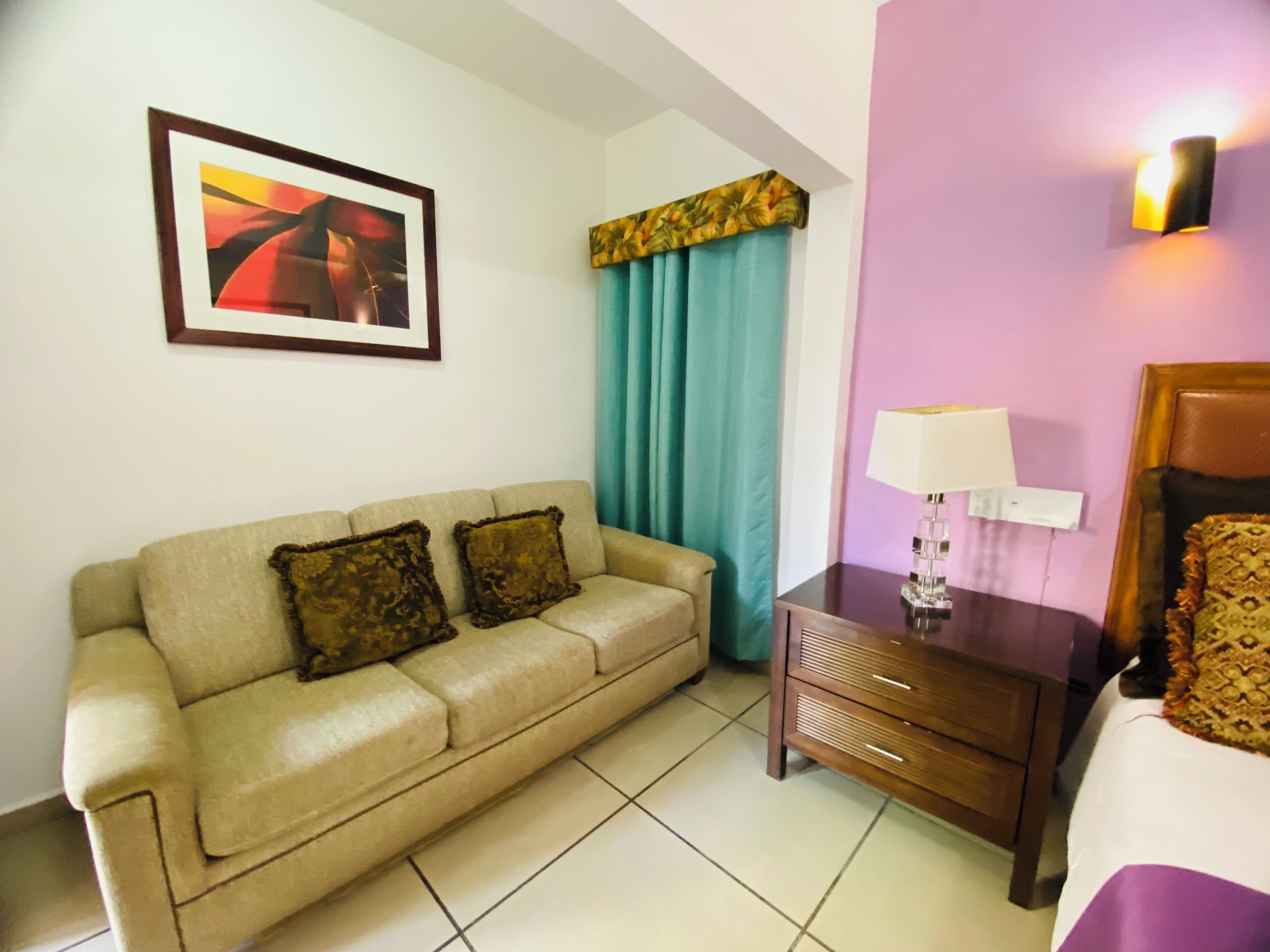 A hotel room with a beige sofa, a small wooden nightstand with a lamp, a bed with a headboard, a window with curtains, and a framed picture on the wall.