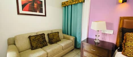 A hotel room with a beige sofa, a small wooden nightstand with a lamp, a bed with a headboard, a window with curtains, and a framed picture on the wall.