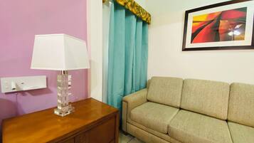 Una habitación con una mesita de noche de madera, una lámpara, un sofá beige, un enchufe en la pared y un cuadro abstracto enmarcado.