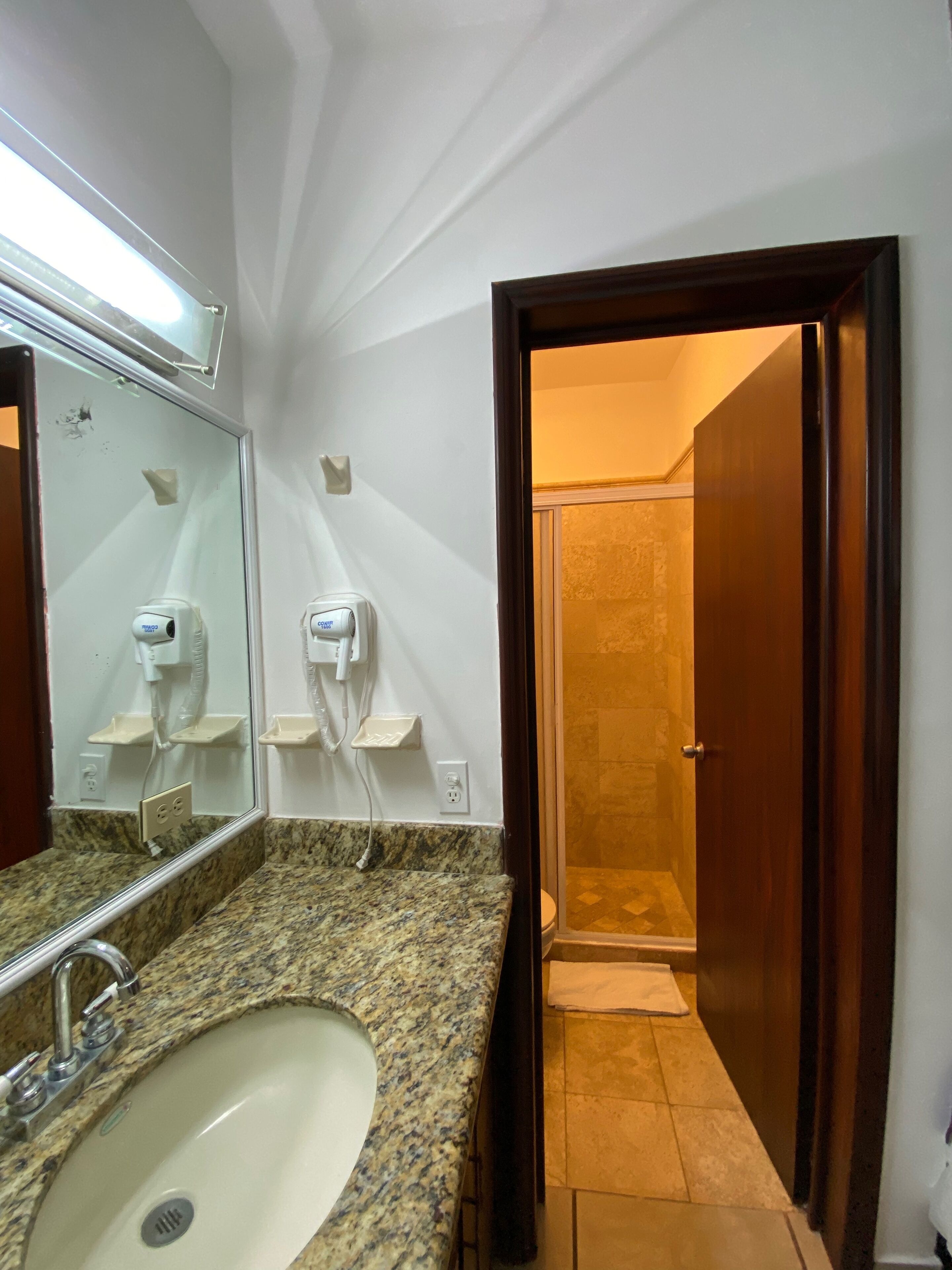 Un baño con lavamanos de encimera de granito, espejo, secador de pelo y ducha visible a través de una puerta abierta.