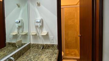 Un baño con lavamanos de encimera de granito, espejo, secador de pelo y ducha visible a través de una puerta abierta.