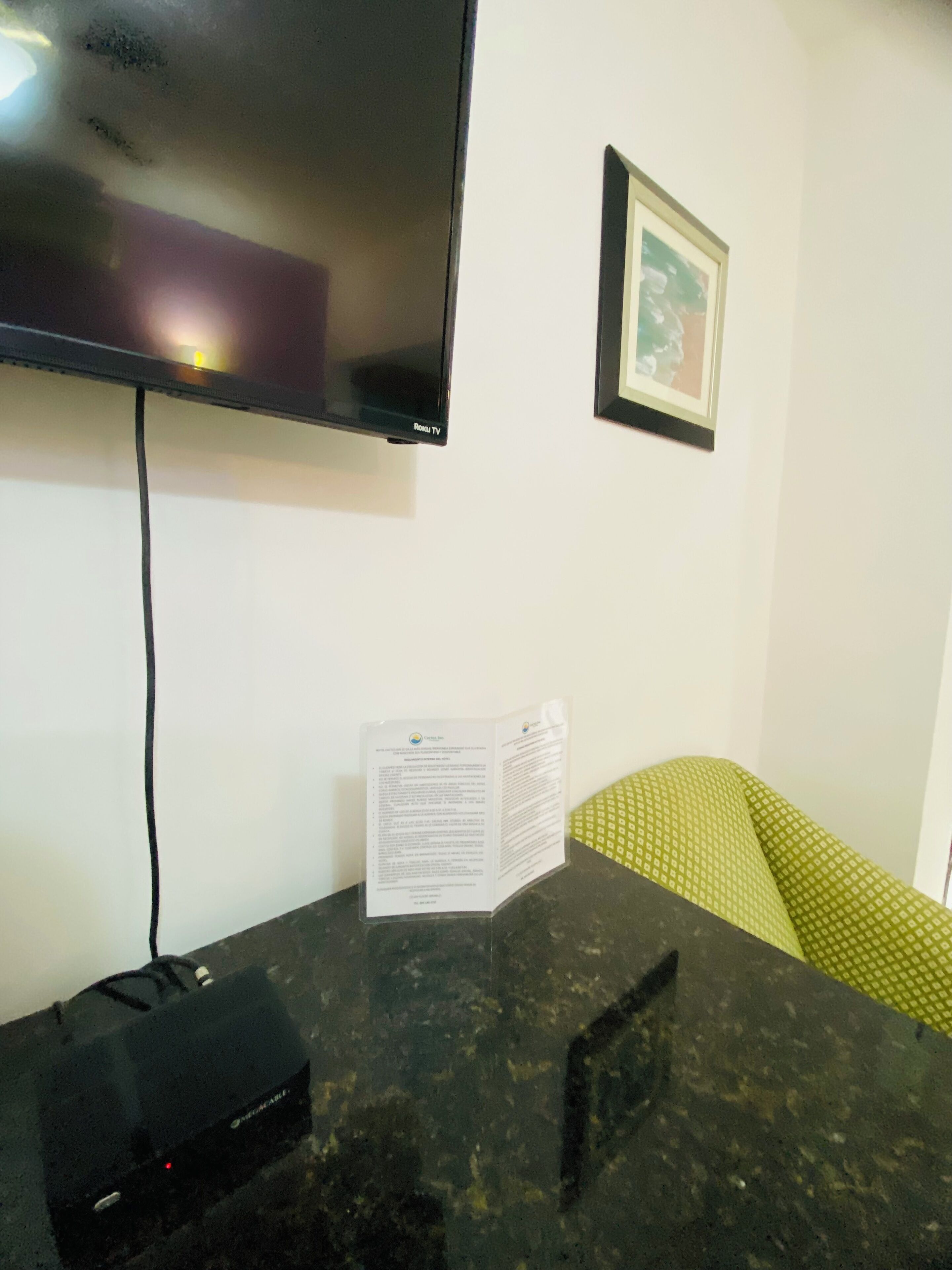 A room with a flat-screen TV mounted on the wall, a table with a power strip, a green patterned cushion, and a framed picture.
