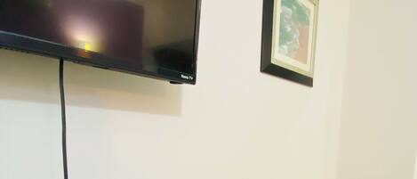 A room with a flat-screen TV mounted on the wall, a table with a power strip, a green patterned cushion, and a framed picture.