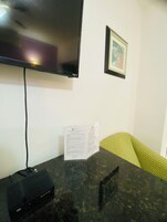 A room with a flat-screen TV mounted on the wall, a table with a power strip, a green patterned cushion, and a framed picture.