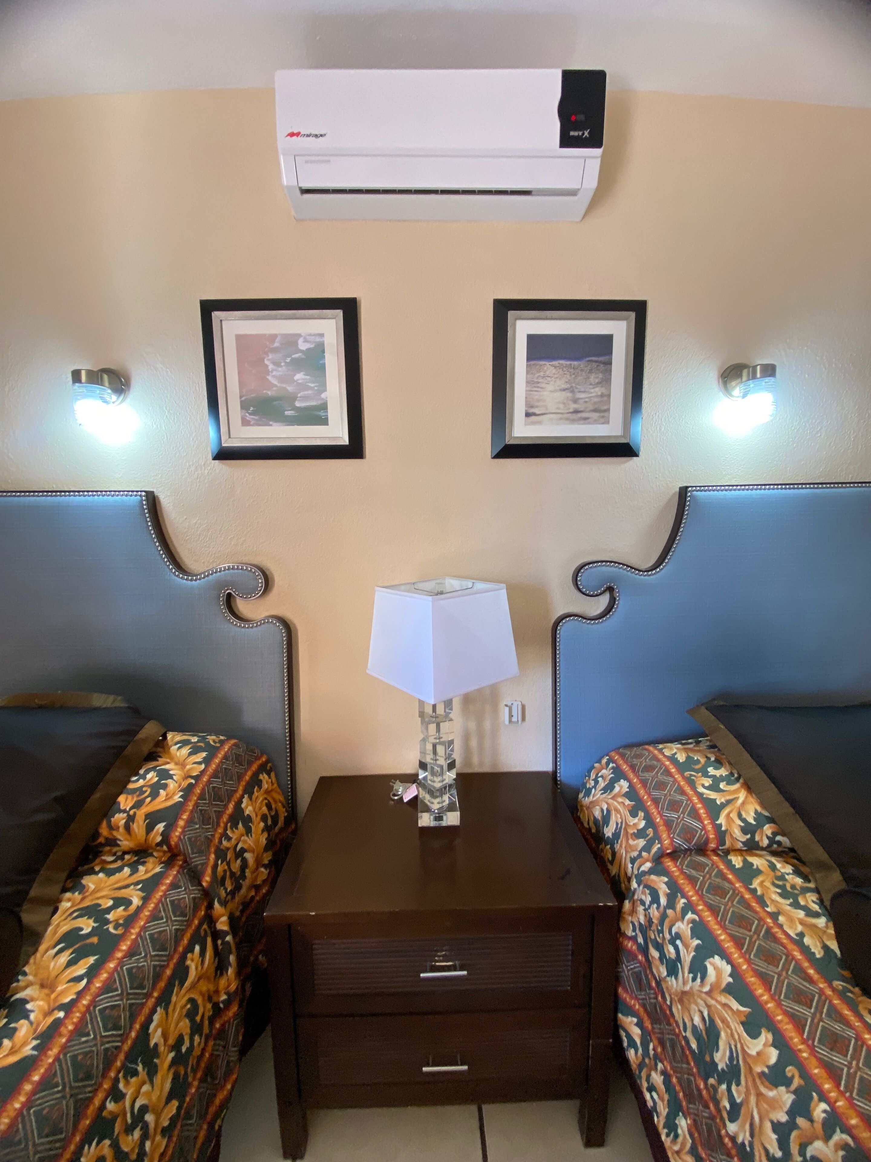 A hotel room with two beds, a nightstand with a lamp, and an air conditioner on the wall.