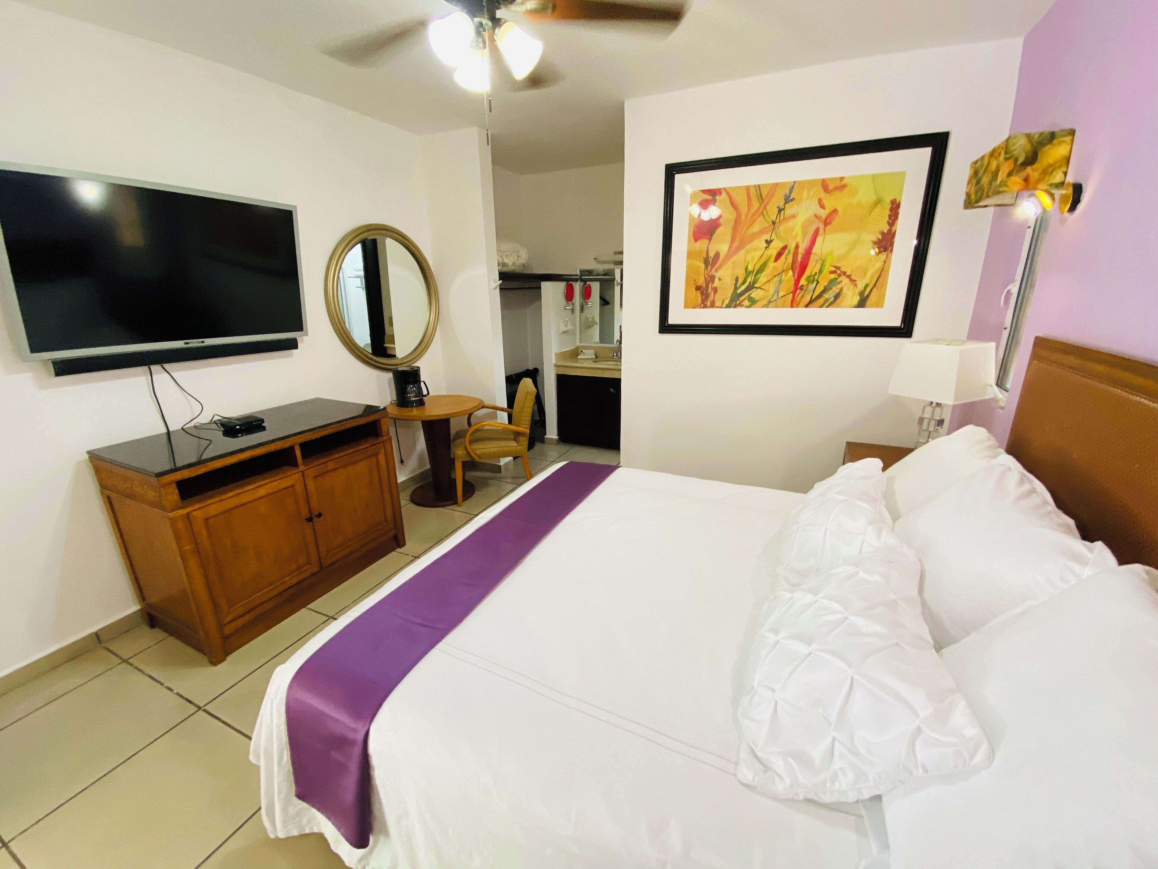 A hotel room with a bed, a television, a desk with a computer, a chair, a mirror, and a painting on the wall.