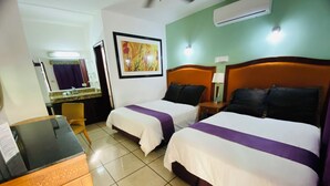 A hotel room with two beds, a desk, a chair, a bathroom with a mirror, and an air conditioning unit.