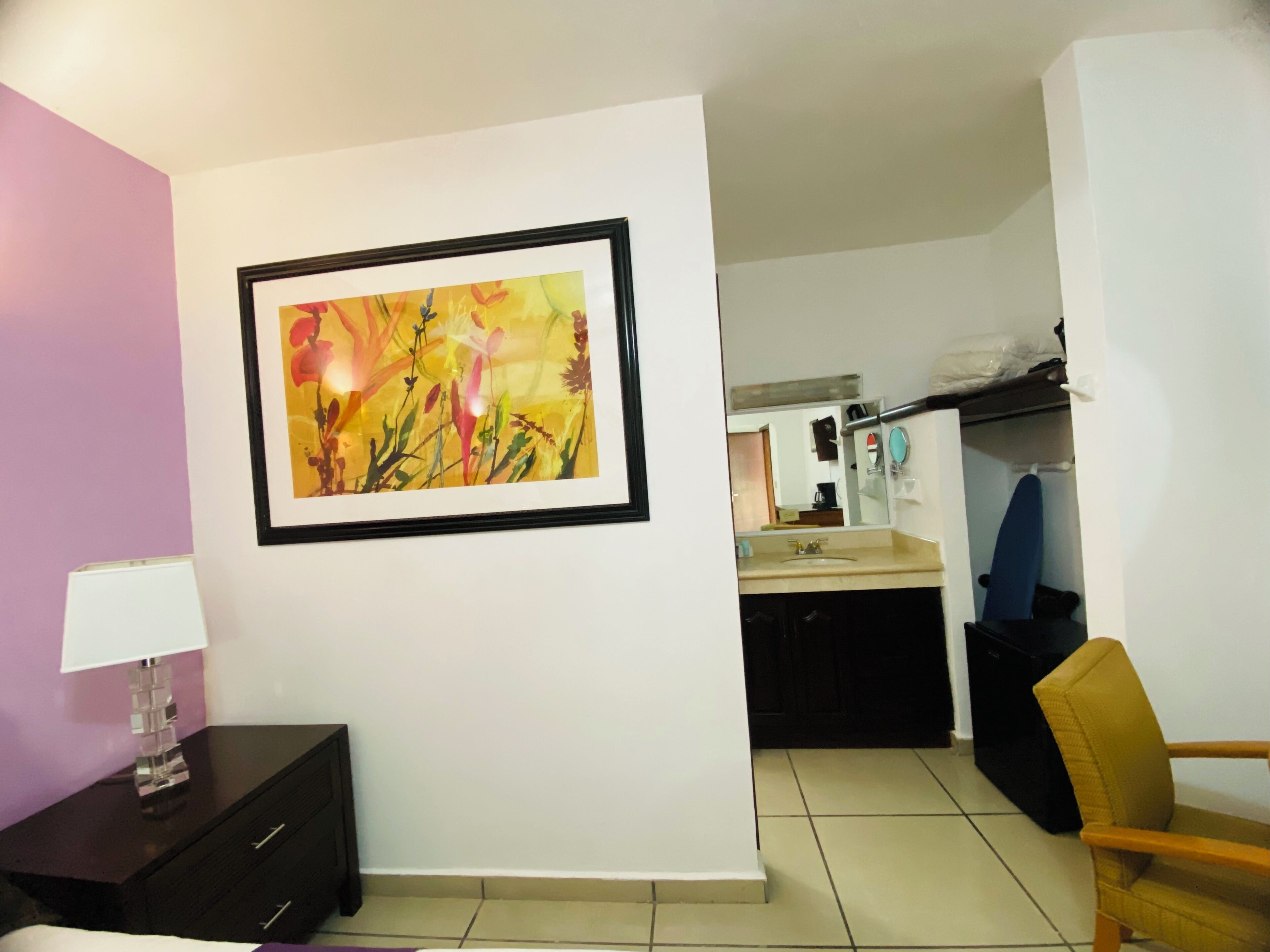 A room with a bed, a bedside table with a lamp, a framed painting on the wall, a chair, and a bathroom visible through an open door.