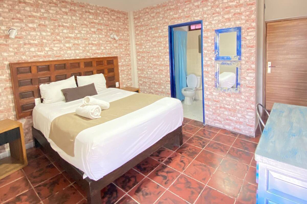 A hotel room with a brick-patterned wall, a wooden bed, a blue-framed mirror, and a tiled floor.
