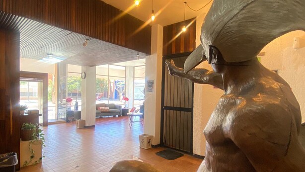 A sculpture of a seated figure in a lobby area with tiled flooring, a wooden ceiling, and hanging lights. - Hotel Flamingos (Guadalajara)