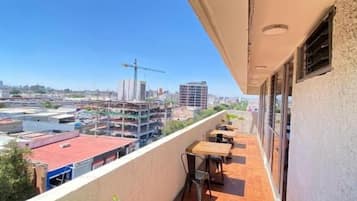 Un balcón con suelo de baldosas, una mesa y sillas, con vistas a un paisaje urbano con edificios y una grúa de construcción.