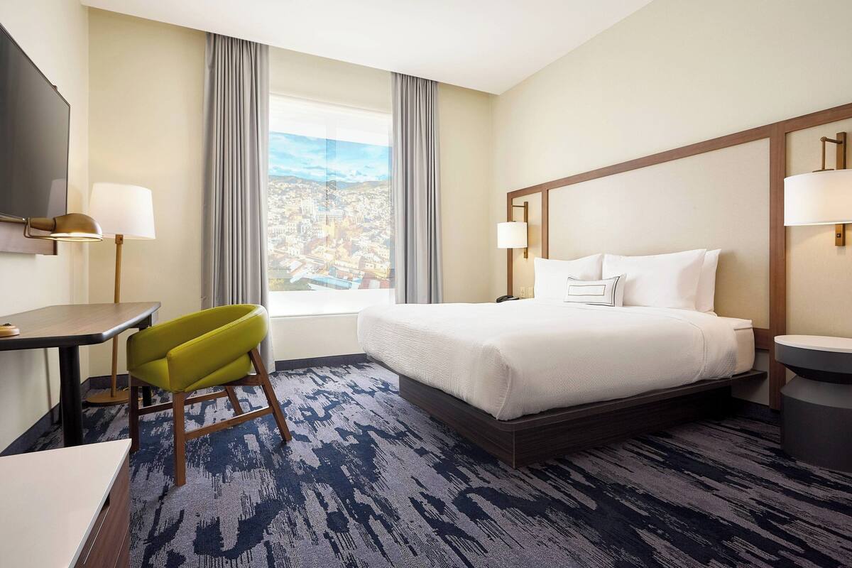 Habitación de hotel con cama, escritorio, silla, televisión y ventana con vista a la ciudad.