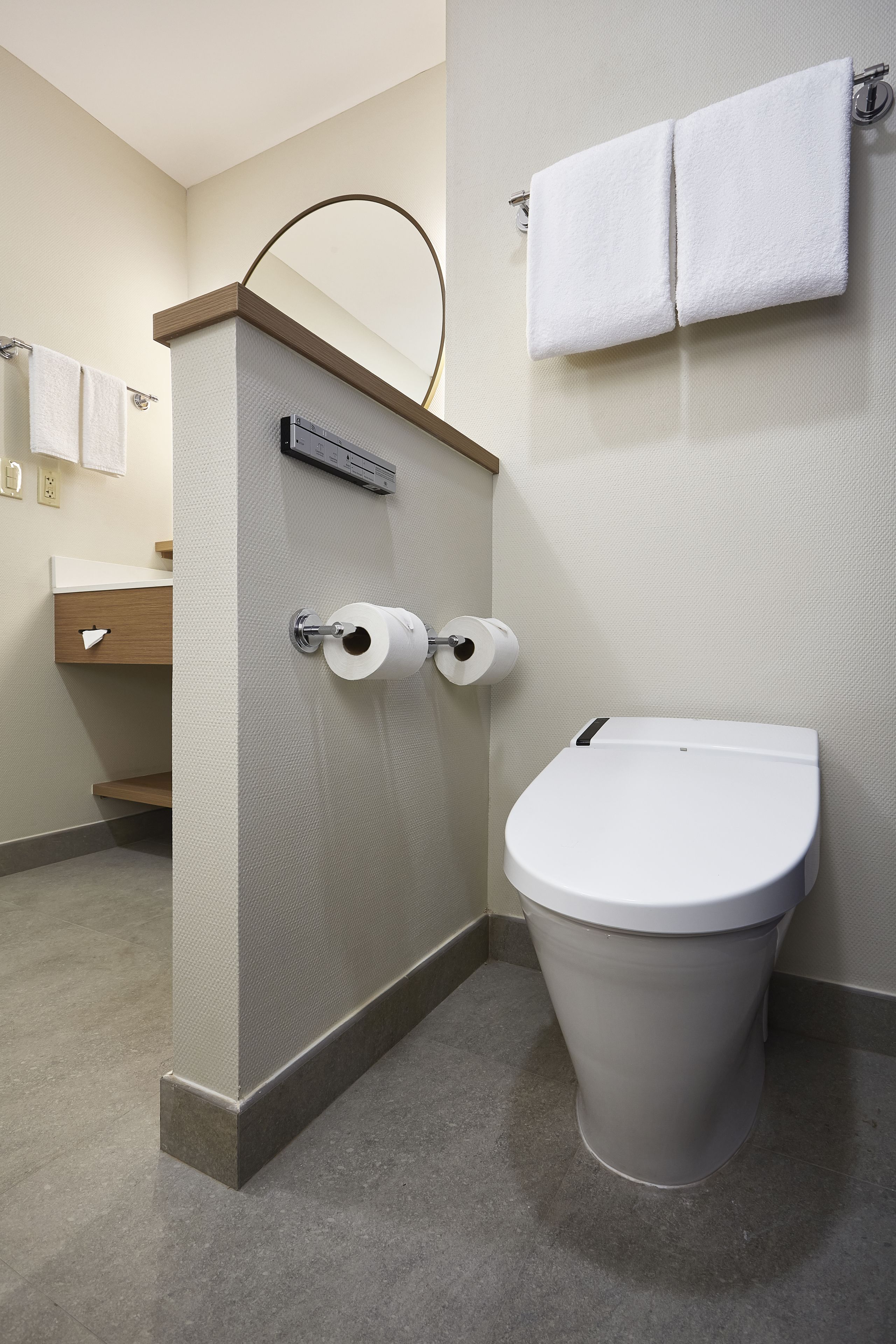 Un baño moderno con un inodoro, dos rollos de papel higiénico, un perchero con dos toallas y un mueble de tocador de madera con espejo.