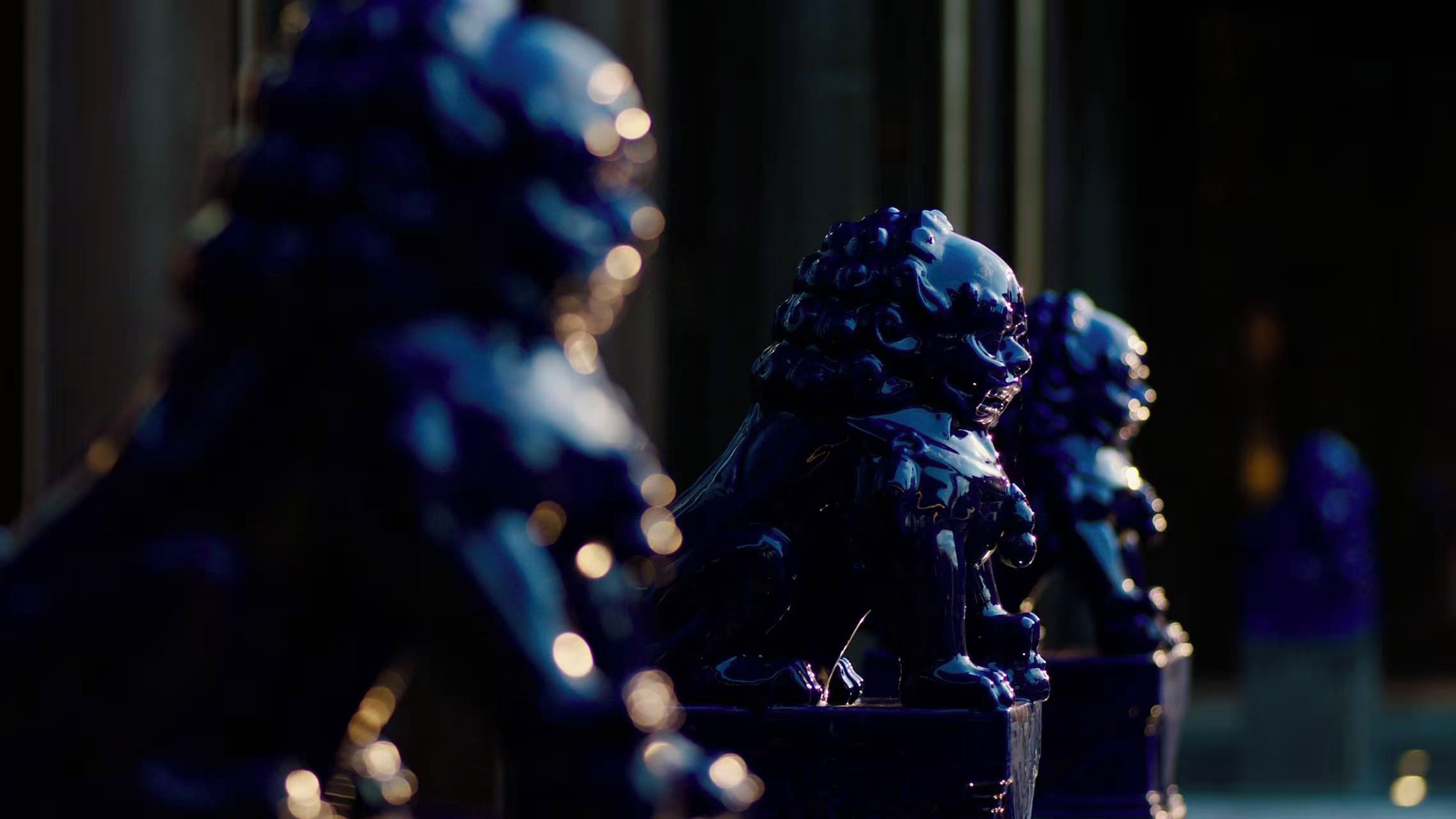 A series of dark-toned statues with a reflective surface, possibly made of metal, positioned against a backdrop with bokeh light effects.