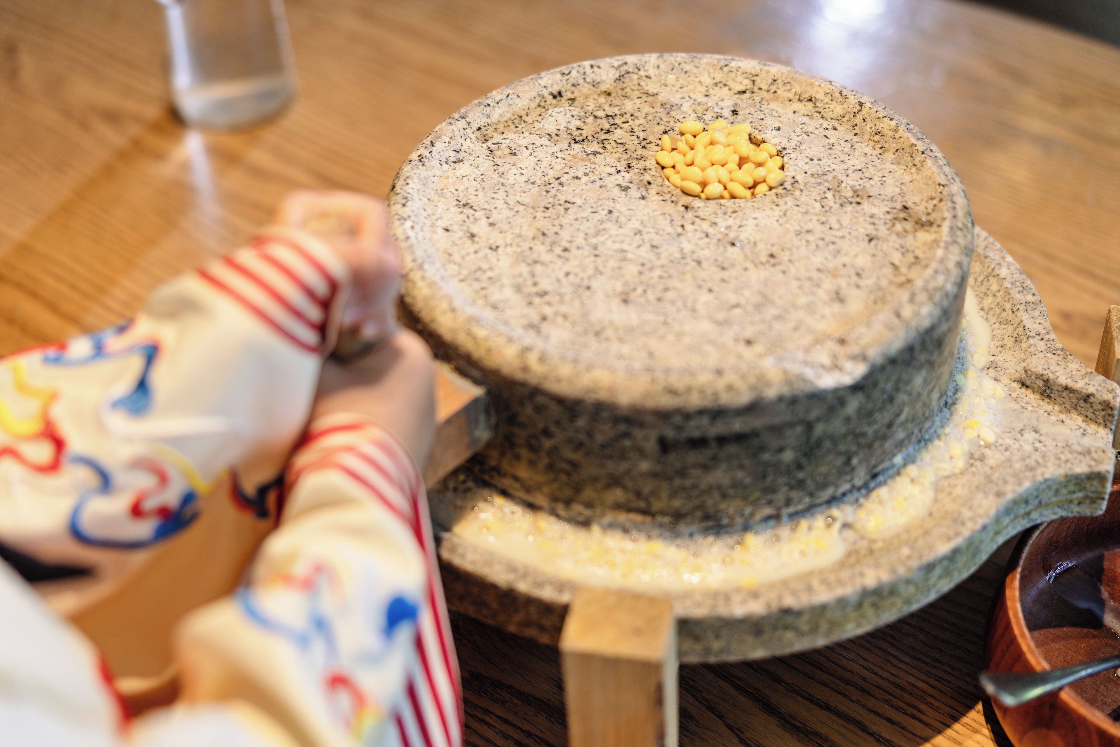A stone mill with a wooden handle, a wooden base, and a metal bowl on a wooden table.