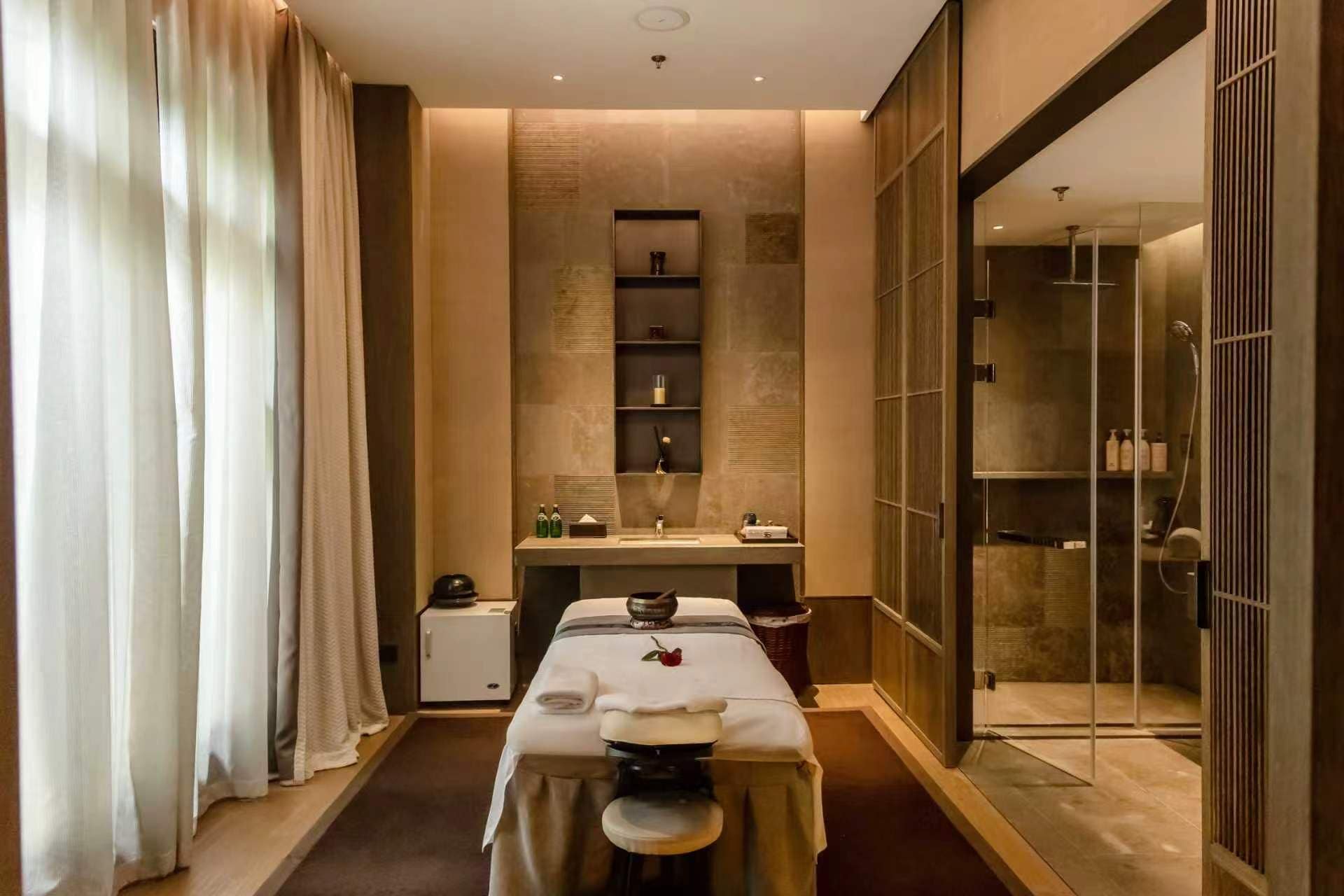 A spa room with a massage table, shelves with toiletries, and a glass shower enclosure.