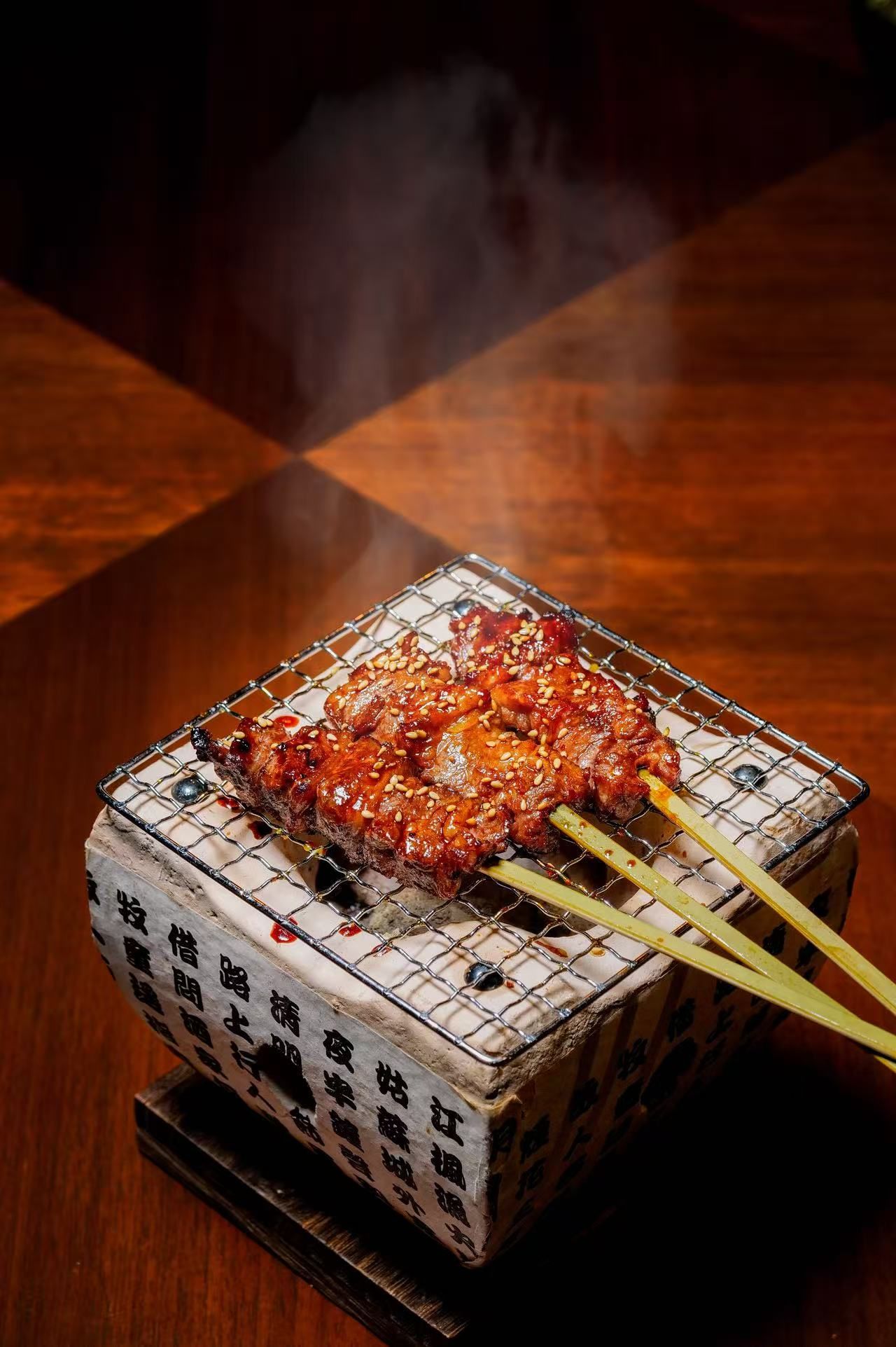 Una parrilla cuadrada con un trozo de carne en brochetas, colocada sobre una mesa con superficie estampada.