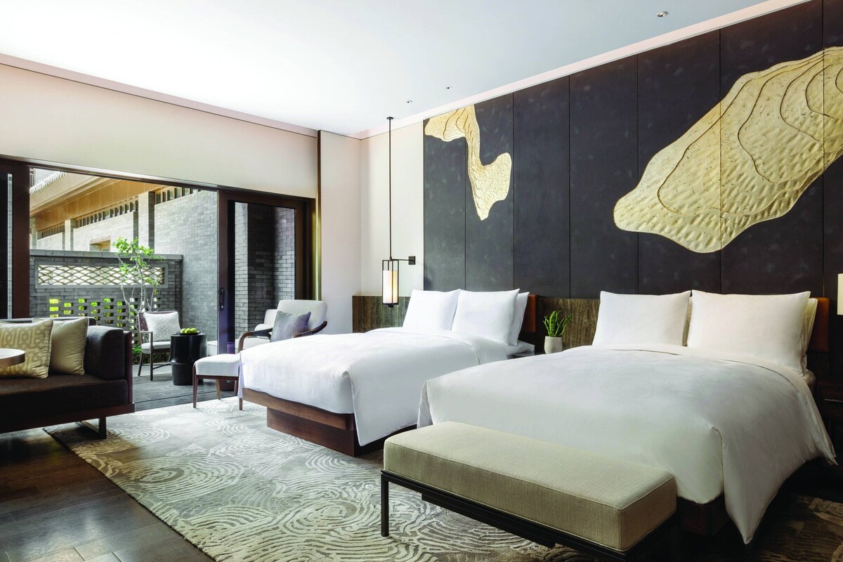 A modern hotel room with two beds, a sofa, and a large window.