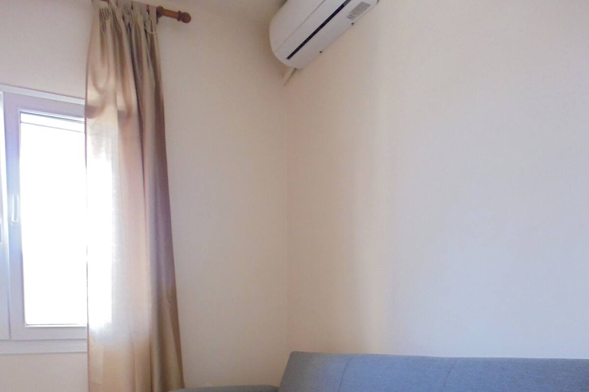 A room with a grey sofa, a window with curtains, and an air conditioning unit on the wall.